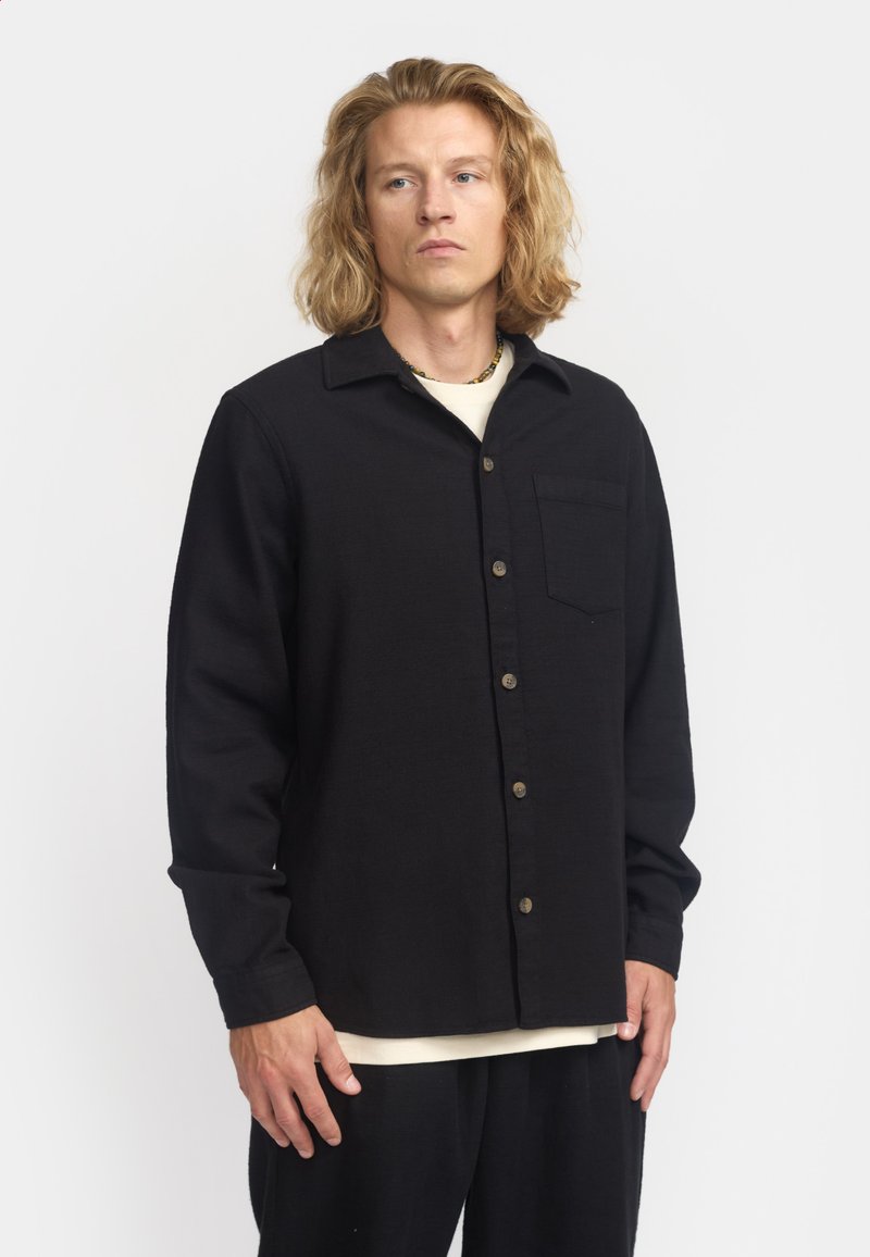 Camisa de botón negra con cuello, mangas largas y un bolsillo en el pecho. Tejido texturizado con botones visibles en un tono contraste.