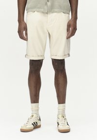 Beige bomullsshorts med manchetter, två framfickor, parat med vita strumpor och krämfärgade sneakers med svarta ränder och guldackcenter.