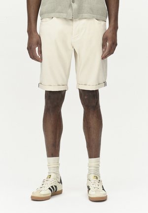 Beige katoenen shorts met omslagen, twee voorzakken, gecombineerd met witte sokken en crèmekleurige sneakers met zwarte strepen en gouden accenten.