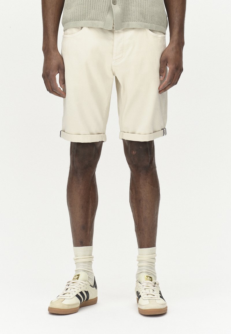 Beige Baumwollshorts mit Bündchen, zwei frontalen Taschen, kombiniert mit weißen Socken und cremefarbene Sneakers mit schwarzen Streifen und goldenen Akzenten.