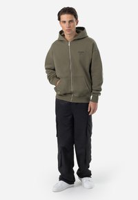 Olive green zip-up hoodie with a kangaroo pocket, paired with black cargo pants featuring side pockets and white sneakers. Simple, casual design.