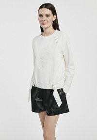 Biała bluza z teksturowanym kwiatowym wzorem, z prążkowanym dekoltem oraz regulowanymi trokami w talii, zestawiona z czarnymi skórzanymi szortami.