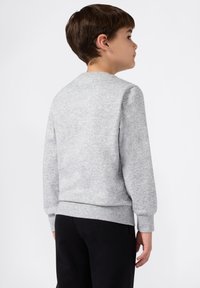 Jongen gezien vanuit de achterkant, gekleed in een lichtgrijze sweatshirt met lange mouwen en zwarte broek, staand tegen een effen witte achtergrond.