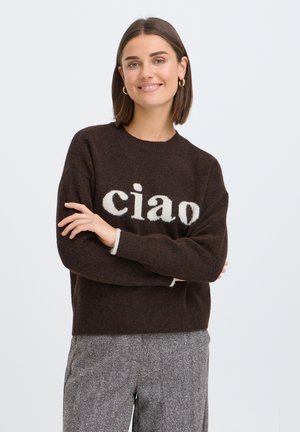 Jeune femme aux cheveux courts bruns portant des boucles d'oreilles créoles dorées, un pull marron foncé avec le texte "ciao" et un pantalon gris, bras croisés et souriante.