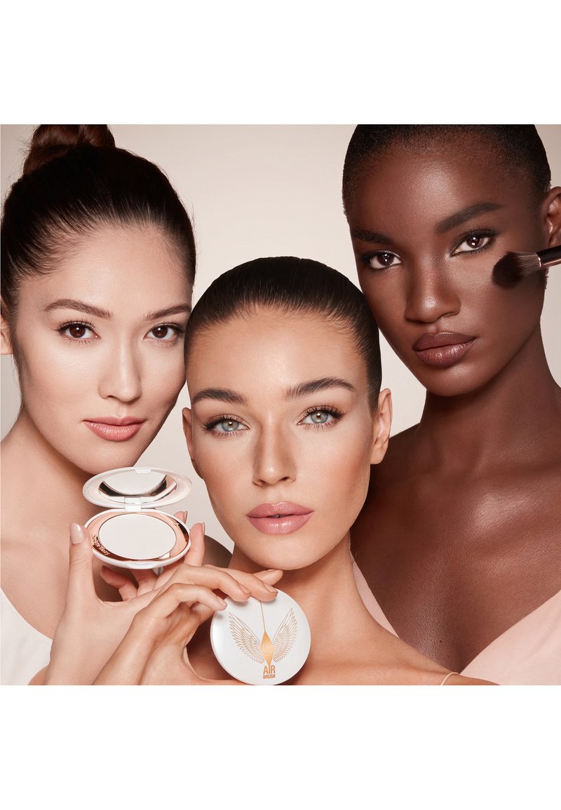 Three women with varied skin tones pose closely; one holds an open compact mirror, another holds a closed compact, and the third applies makeup with a brush.
