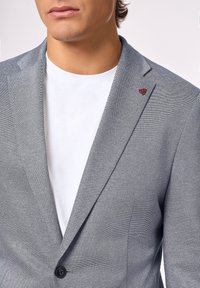 Blazer grigio chiaro con trama testurizzata, chiusura con un singolo bottone nero e accento rosso sulla patta, indossato sopra una t-shirt bianca.