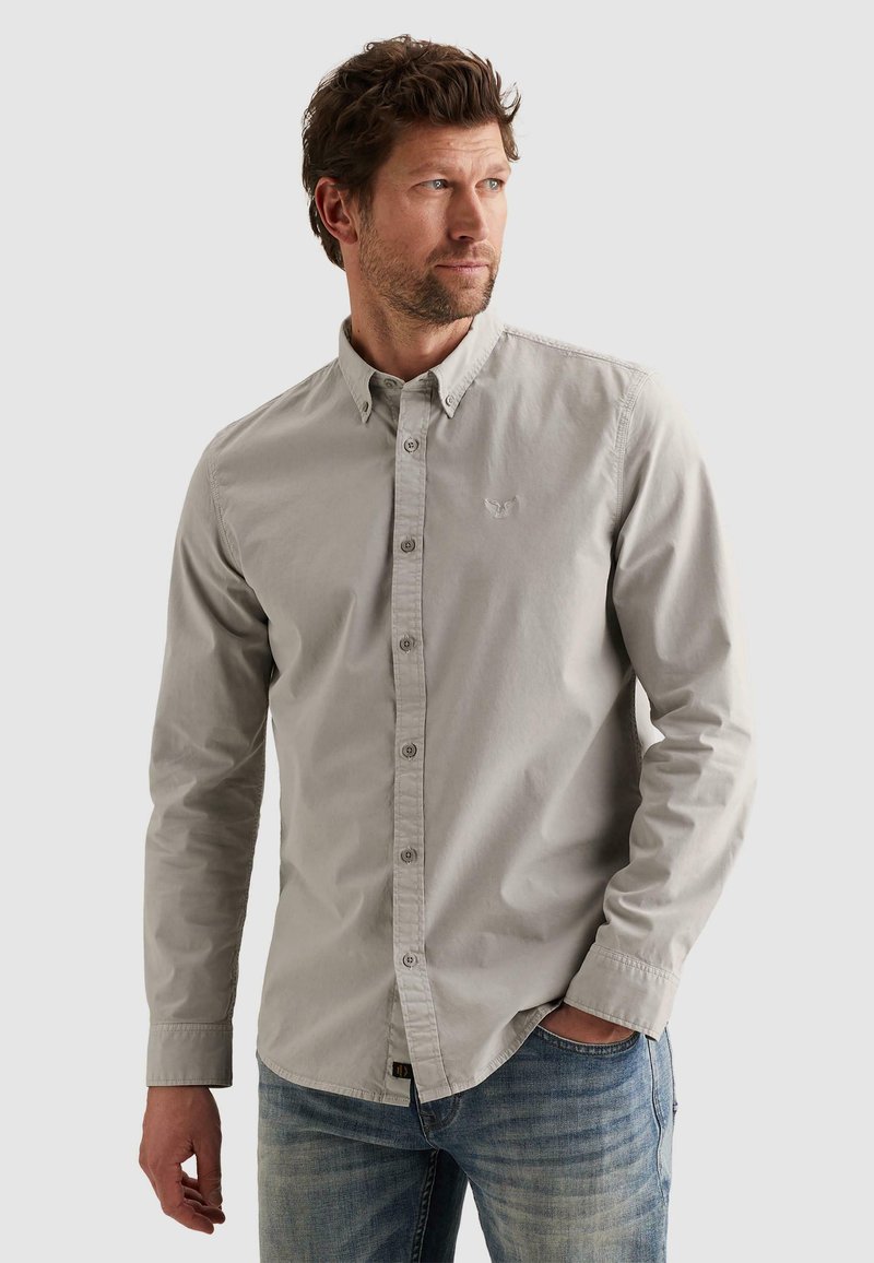 Muž s krátkymi hnedými vlasmi, oblečený v svetlosivej košeli na gombíky a modrých džínsoch, pozerá sa doľava s jednou rukou v vrecku.