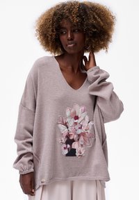 Vrouw met krullend haar die een losse mauve sweater draagt met een bloemen- en vlinderontwerp over een lichte rok op een witte achtergrond.