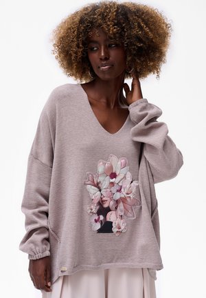 Femme avec des cheveux bouclés portant un pull mauve ample orné d'un motif floral et de papillons sur une jupe claire, sur fond blanc.
