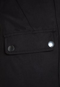 Detalle de chaqueta de tela negra que presenta un bolsillo rectangular con cierre a presión, superficie texturizada y costuras visibles.