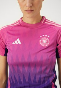 Maillot de soccer rose et violet avec des motifs triangulaires, logo Adidas blanc et emblème d'équipe circulaire avec des étoiles et du texte.