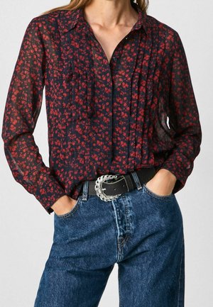 Vrouw draagt een marineblouse met kleine rode bloemenprint, ingestopt in een blauwe spijkerbroek, geaccentueerd met een zwarte riem met een zilveren gesp.