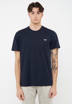 Jonge man met kort krullend haar draagt een navy blauw t-shirt met logo op de borst en licht beige broek, toont tatoeages op de rechter onderarm.
