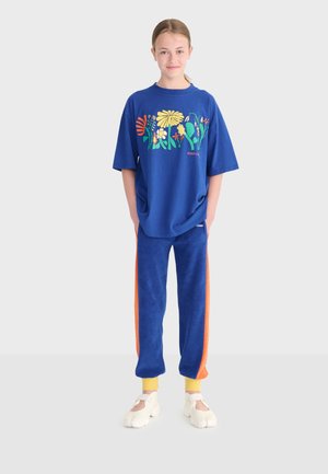 Fille debout portant un T-shirt bleu surdimensionné à imprimé floral, un pantalon de survêtement bleu avec des rayures orange, des chaussettes jaunes et des chaussures blanches à bout fendu.