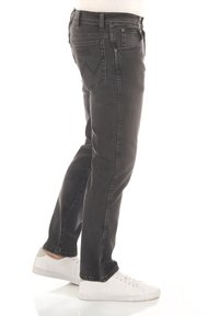 Jeans negros de corte slim con diseño de cinco bolsillos, costuras visibles y cierre con cremallera. El modelo lleva zapatillas blancas y está de perfil.