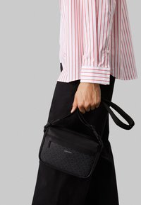 Sort crossover taske med tekstureret front, der har et logo. Den har en aftagelig rem og en rektangulær facon. Bæres sammen med en hvid og rød stribet skjorte.