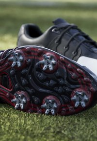Black and white golf shoes resting on grass, with cleated soles visible showing six soft spikes on the bottom.