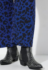 Zwarte enkellaarzen met zilveren studs, met een lage hak en zij-elastische panelen, gecombineerd met blauwe broeken met een zwart luipaardprint.
