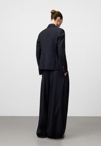 Donna con i capelli raccolti a chignon, indossante un blazer blu scuro e pantaloni ampi, in piedi con le mani in tasca davanti a uno sfondo neutro.