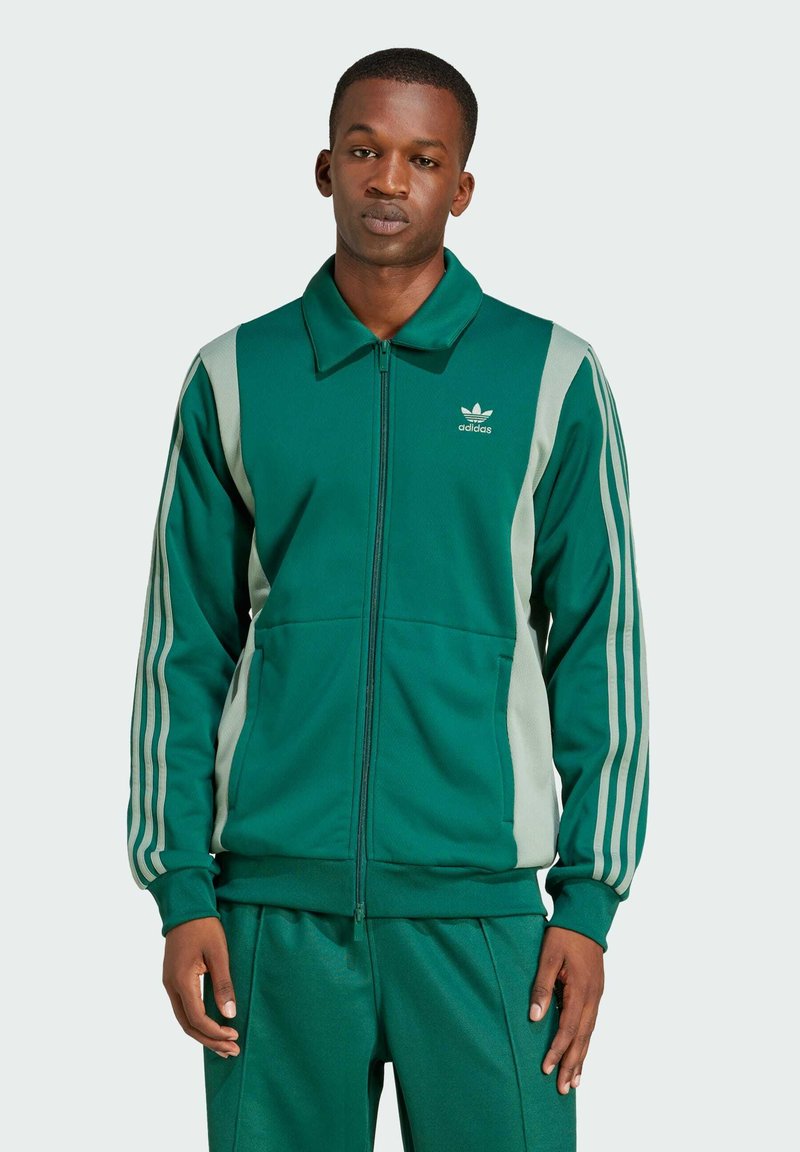 Zeleni zip-up sportski jakna s teksturiranim završetkom, koja ima sive bočne panele i tri bijele pruge duž rukava. Logo je prikazan na prsima.