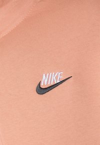 Persikofärgat tyg med en mjuk textur och en kontrasterande svartvit broderad Nike-logotyp på framsidan.