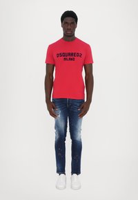 Camiseta de algodón roja con "DSQUARED2 MILANO" impreso en negro; combinada con jeans azules desgastados y zapatillas blancas, de pie contra un fondo blanco.