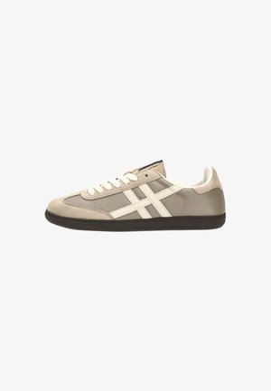 Zapatilla deportiva de color beige claro con acentos blancos, materiales de ante y malla, puntera redonda, suela plana de goma negra y diseño de rayas en contraste.