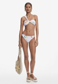Bikini set s dizajnom na jednom ramene, bielym základom a farebnými vzormi mušlí. Modelka drží tkanú tašku s detailmi hviezdoviek. Ľahká obuv.