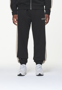 Pantalones de chándal negros con rayas laterales beige, puños elásticos y un detalle de texto con el logo. Llevados con zapatillas deportivas blancas.