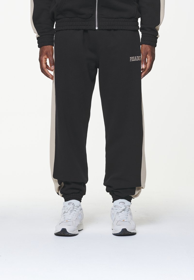 Pantalones de chándal negros con rayas laterales beige, puños elásticos y un detalle de texto con el logo. Llevados con zapatillas deportivas blancas.
