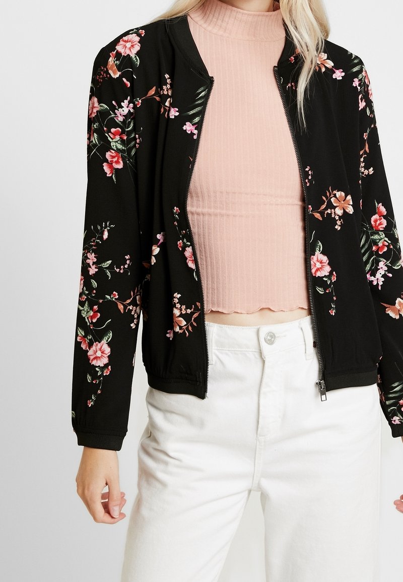 Femme portant une veste zippée noire à imprimé floral sur un crop top côtelé rose et un pantalon blanc taille haute, mains détendues sur les côtés.