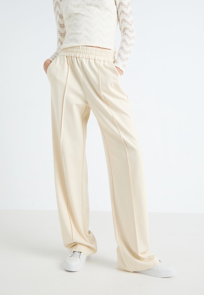 Osoba v krémových širokých kalhotách s elastickým pasem, rukama v kapsách, v bílých teniskách a dlouhém krémovém krajkovém topu s dlouhými rukávy.