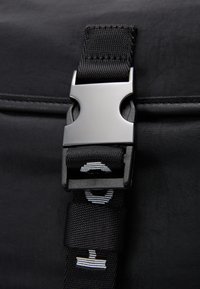 Cinghia di un bagaglio in tessuto nero con un fibbia metallica argentea e nastro nero con dettagli a strisce bianche. La vista ravvicinata mette in evidenza la texture e i componenti metallici.