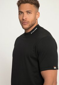 Camiseta negra de manga corta con un cuello alto. Presenta un collar de canalé con el logo "STHUGE" y una etiqueta naranja en la manga izquierda.