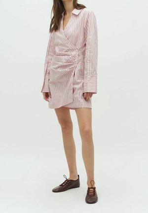 Kobieta ubrana w różowo-białą sukienkę kopertową w pionowe paski z długimi rękawami oraz brązowe płaskie buty sznurowane, stojąca na białym tle.