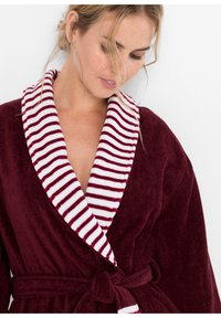 Accappatoio bordeaux con colletto a righe bianche e rosse, cintura e texture morbida. Il tessuto appare morbido e accogliente.
