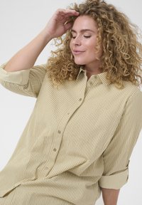 Frau mit lockigem blondem Haar, die ein beigefarbenes gestreiftes Hemd mit Knöpfen trägt, berührt mit einer Hand die Stirn, die Augen geschlossen und lächelt leicht.