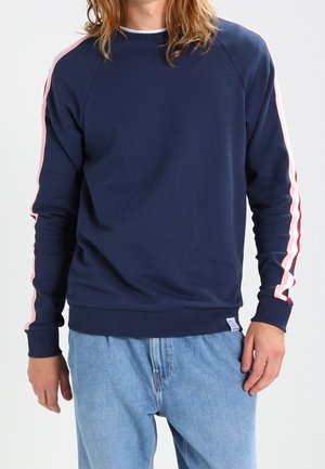 Osoba ubrana w granatową bluzę z różowymi paskami na rękawach i jasnoniebieskie jeansy, stojąca na jednolitym tle.