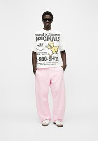 Άνδρας που φοράει μαύρα γυαλιά ηλίου, λευκό T-shirt Adidas Originals με γραφικό κινουμένων σχεδίων πάπιας, χαλαρά ροζ παντελόνια και λευκά αθλητικά παπούτσια, στέκεται μπροστά σε λευκό φόντο.