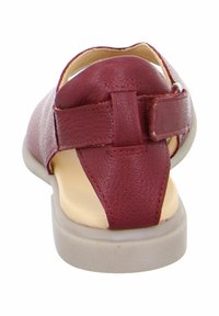 Red leather sandal with a textured upper, back strap, and flat, light-colored sole. Features a hook-and-loop closure for adjustable fit.