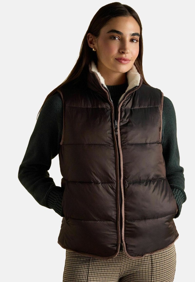 Hnědá vycpaná vesta se zipovým zapínáním a smetanovým límcem. Hladká textura, boční kapsy a kontrastní lemování podél okrajů.
