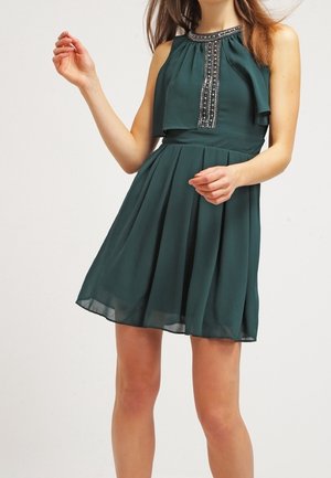 Woman wearing a sleeveless dark green dress with decorative beading on the neckline, standing against a plain white background.