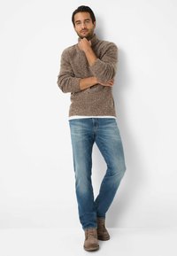 Bruine gebreide coltrui van textuurstof, lichtblauwe spijkerbroek en bruine enkelboots. Model staat met gekruiste armen.