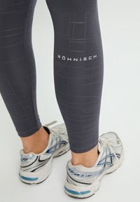 Leggings atléticos grises con un sutil patrón geométrico, que presentan un logo blanco. De longitud hasta el tobillo y con una textura suave y mate.