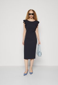 Mornarsko modra midi obleka z naboranimi detajli na ramilih, v kombinaciji s svetlomodrimi slingback petami in usklajeno torbico. Model nosi sončna očala.