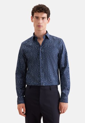Mann mit kurzen lockigen Haaren, der ein dunkelblaues langärmliges Hemd mit dezentem Blumenmuster und schwarze Hose trägt.