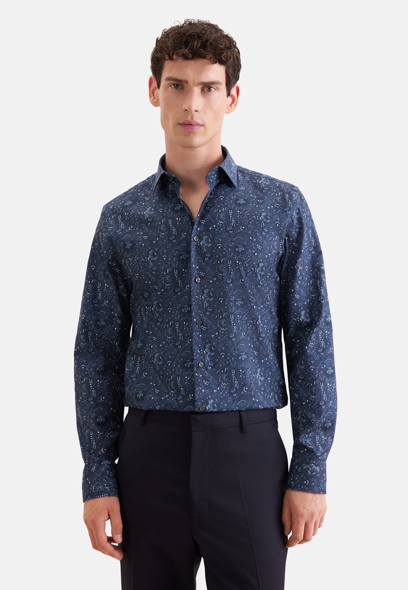 Homme aux cheveux courts et bouclés portant une chemise bleu foncé à manches longues avec un motif floral subtil et un pantalon noir.