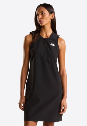 Femme portant une robe noire sans manches avec une fermeture éclair à l'avant et le logo The North Face sur la poitrine, debout devant un fond blanc uni.