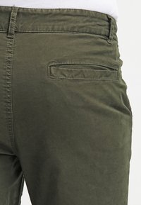 Olivegröna bomullsbyxor med ett sydd midjeband, bakficka med klaff och en slät, något texturerad yta.