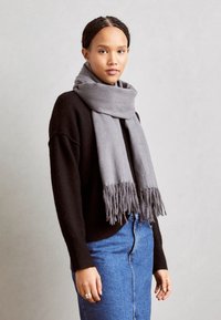 Écharpe grise à franges, en matière douce, portée sur un pull noir, associée à une jupe en denim. Fond neutre.